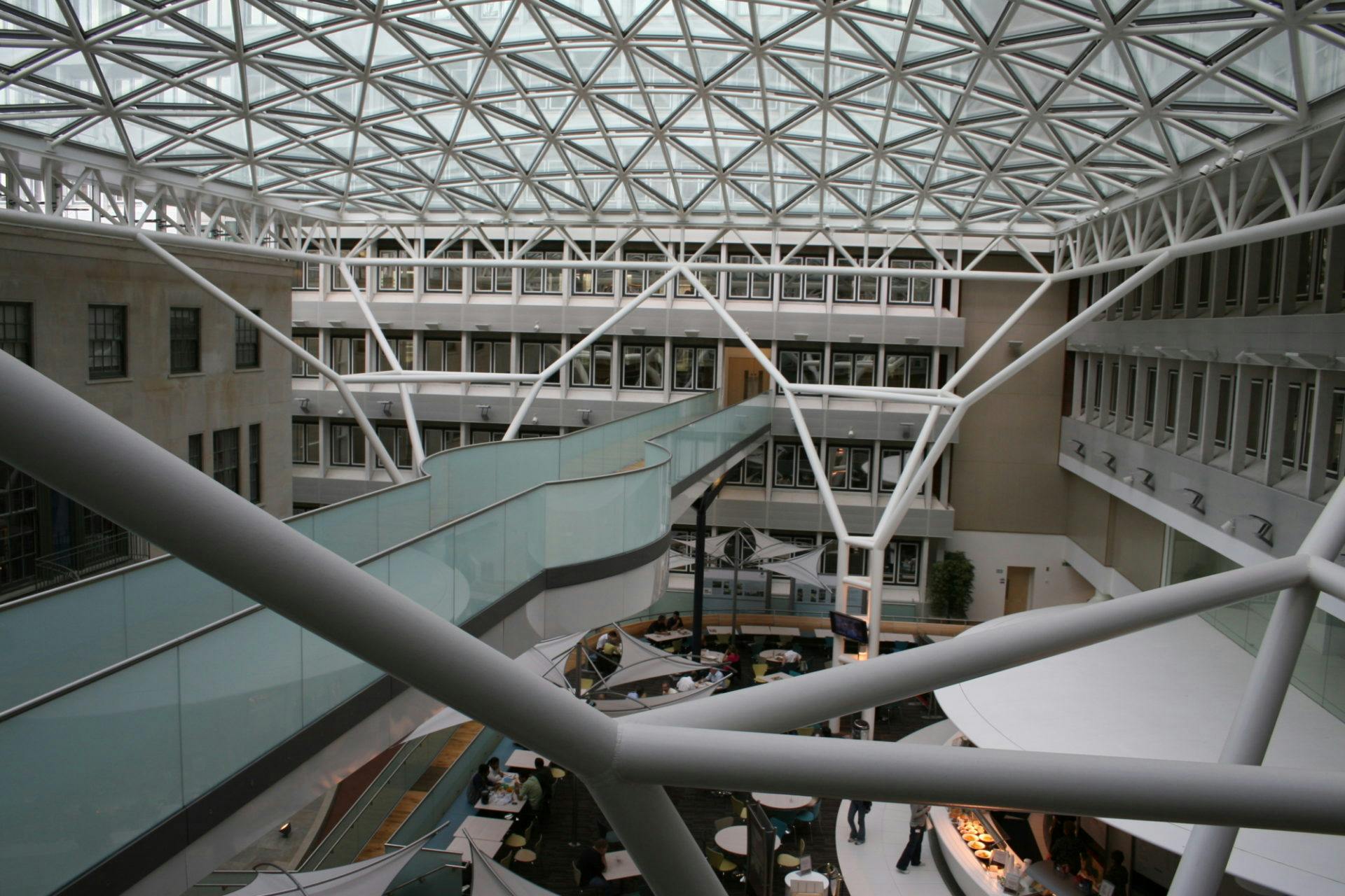 Interior of a modern building with a geometric glass ceiling, featuring multiple levels, a central café area, and intersecting white beams.